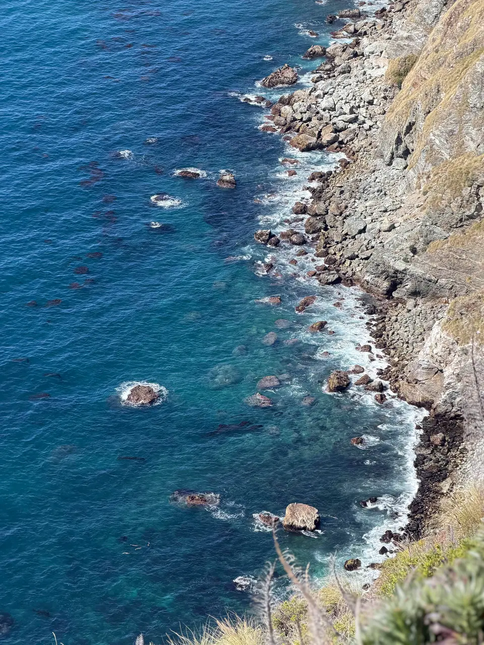 Looking down a rocky Big Sur shoreline where deep blue and turquoise water meets boulders and cliff.